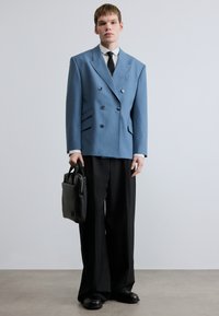 Sacou bleumarin cu dungi fine, cu două rânduri de nasturi argintii, asortat cu pantaloni negri largi și ținând o geantă neagră cu textură.