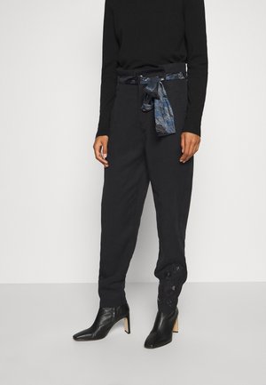 Personne portant un pantalon noir taille haute avec une ceinture en tissu bleu à motif, un haut noir à manches longues et des bottines noires à talons.