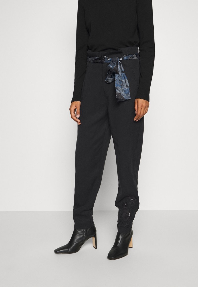 Personne portant un pantalon noir taille haute avec une ceinture en tissu bleu à motif, un haut noir à manches longues et des bottines noires à talons.
