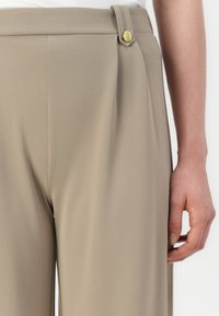Beige tailored pants in a textured fabric, featuring a side button detail and a slim fit. Close-up of the waist and hand holding the fabric.