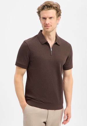 Mann trägt dunkelbraunes, strukturiertes Kurzarm-Poloshirt mit Reißverschluss, beige Hose, steht mit einer Hand in der Tasche und blickt leicht zur Seite.