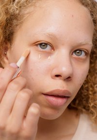 Concealer applicator held near the eye, creamy beige product on skin, close-up of light skin tone and textured, wavy blonde hair.
