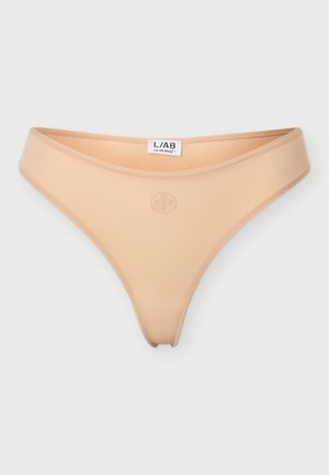 Mujer de pie descalza vistiendo un conjunto de ropa interior beige sin costuras sobre un fondo liso y claro.
