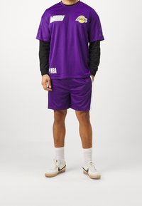 Mężczyzna ubrany w fioletowy strój koszykarski Los Angeles Lakers z czarnymi długimi rękawami, białe skarpetki i białe buty sportowe Nike.