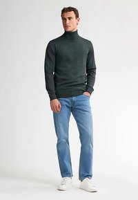 Donkergroene coltrui in een aansluitend model, gecombineerd met lichtblauwe spijkerbroek en witte sneakers. Eenvoudige en minimalistische stijl.