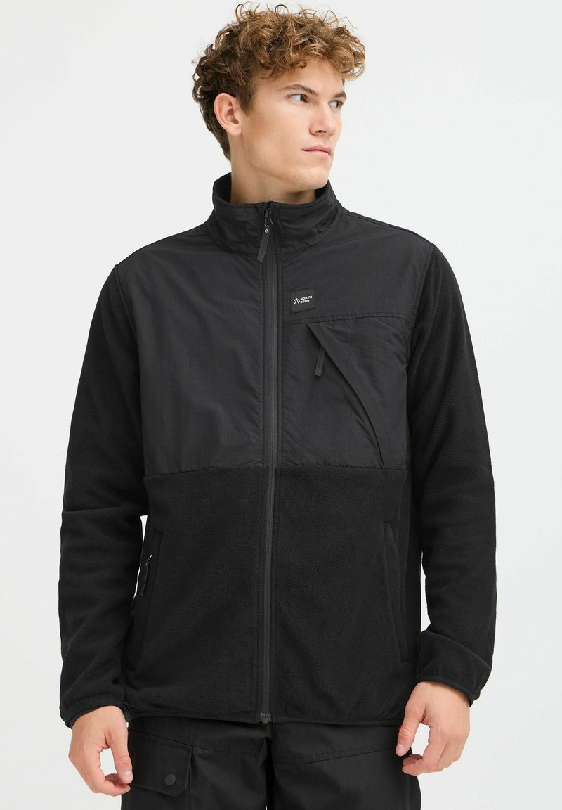 Svart jakke laget av en blanding av glatt, lett stoff og strukturert fleece. Har høy krage, glidelås foran og lomme.