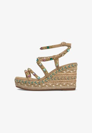 Sandalia de cuña beige con plataforma de yute trenzado, adornos de gemas multicolores y tachuelas doradas, y correa ajustable en el tobillo sobre fondo blanco.