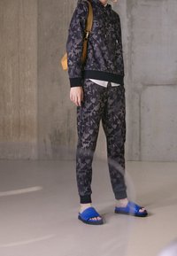 Felpa nera mimetica con tasca a marsupio, abbinata a pantaloni joggers coordinati e ciabatte blu. L'outfit presenta polsini a coste.
