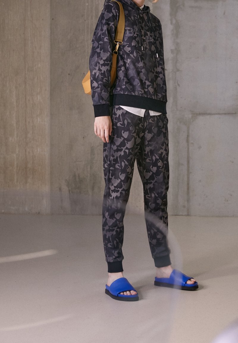 Felpa nera mimetica con tasca a marsupio, abbinata a pantaloni joggers coordinati e ciabatte blu. L'outfit presenta polsini a coste.