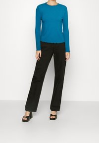 Blå randig långärmad topp med vågig kant, matchad med svarta vida jeans och svarta klacksandaler med blockklack. Enfärgad bakgrund.