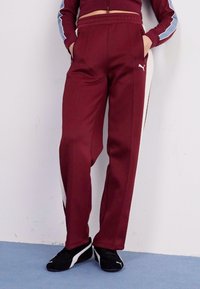 Bordeauxrode trainingsbroek van glad, dik stof met witte zijstreepjes en een elastische tailleband. Gestyled met zwarte sportschoenen.