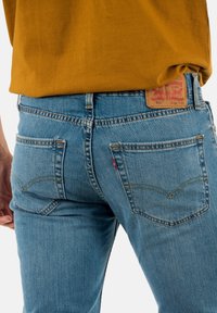 Jean en denim bleu avec une coupe slim, comportant cinq poches, des coutures jaunes, et une étiquette en cuir sur la taille au dos.