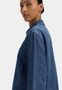 Veste en denim bleu foncé avec un col classique, des manches longues et une coupe décontractée ; présente des coutures visibles et une surface légèrement texturée.