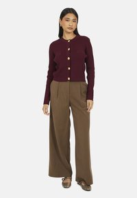 Femme portant un cardigan bordeaux à boutons, un pantalon large marron et des ballerines à motif serpent, se tenant avec une expression neutre.