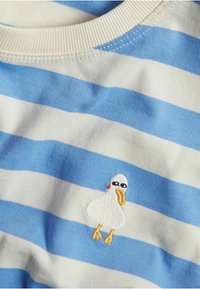 Raidallinen sinivalkoinen puuvillapaita. Vasemmassa rinnassa on brodattu piirrosankka, joka pitää aurinkolaseja ja jolla on keltainen nokka.