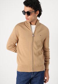 Pull zippé beige fabriqué en maille lisse, avec un col montant, des poignet et un ourlet côtelés, porté par-dessus un pull crème.