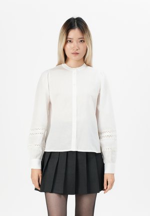 Hvit langermet bluse med knappestenging foran og blonder detaljer på ermene, kombinert med et svart plissert skjørt. Glatt stoffstruktur.