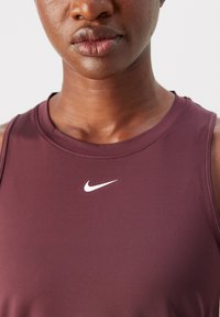Burgundski športni top z gladkim materialom, okroglo izrezano ovratnikom in belim logotipom Nike na prsih. Poudarjen pogled na izrez in teksturo blaga.