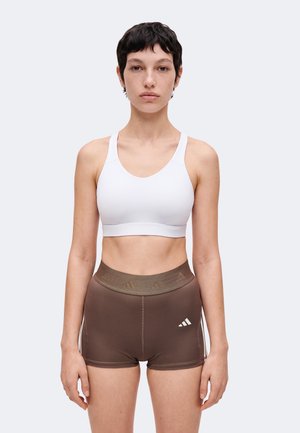 Žena s krátkymi tmavými vlasmi, ktorá nosí bielu športovú podprsenku a hnedé športové šortky s logom Adidas, stojí na svetlom pozadí.