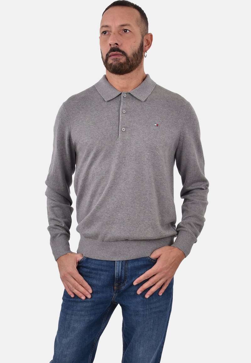 Maglione polo grigio con colletto, placket a tre bottoni, polsini e orlo a costine. Presenta un piccolo logo sul lato sinistro del petto. Indossato con jeans blu.