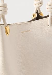 Krémsvá kožená kabelka s uzlovatými uchy a zlatými kovovými doplňky, s vyraženým názvem značky "Coccinelle" ve zlaté barvě.