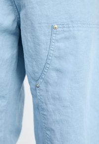 Lichtblauwe stof met een gestructureerd oppervlak, voorzien van een zijzak die is beveiligd met een zilveren drukknop. Eenvoudig ontwerp met zichtbare stitching.
