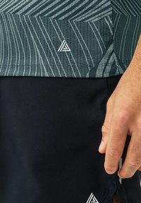 Kortærmet atletisk top i mørkegrøn med et geometrisk mønster og tekstureret stof, parret med sorte shorts. Har små logoakcentre.