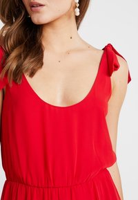 Robe rouge avec un décolleté large et des bretelles à nœud réglables. Fabriquée en tissu lisse, elle présente une taille cintrée et une jupe fluide.
