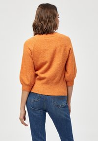 Maglione arancione lavorato a maglia con scollatura rotonda e maniche a sbuffo tre quarti, abbinato a jeans azzurri a vita alta. Tessuto testurizzato.