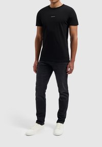 Zwarte katoenen t-shirt met een ronde halslijn, voorzien van een klein wit logo op de borst. Gecombineerd met donkere denim jeans en witte sneakers.