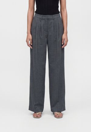 Donna che indossa pantaloni grigi a vita alta e gamba larga e sandali beige, in piedi davanti a uno sfondo chiaro e uniforme.