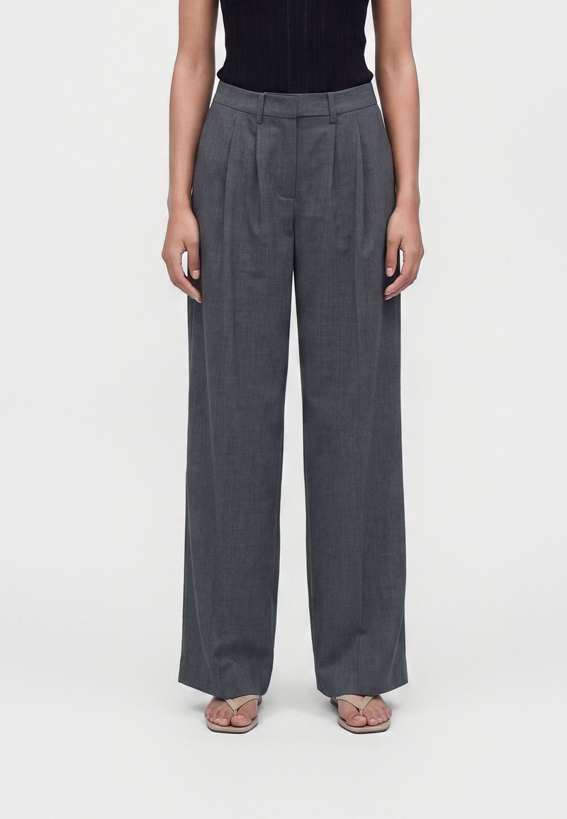 Femme portant un pantalon gris taille haute à jambes larges et des sandales beiges, debout devant un fond clair uni.