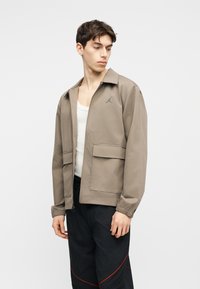 Giovane uomo che indossa una giacca beige con zip sopra una canottiera bianca e pantaloni neri con dettagli rossi su uno sfondo chiaro e uniforme.