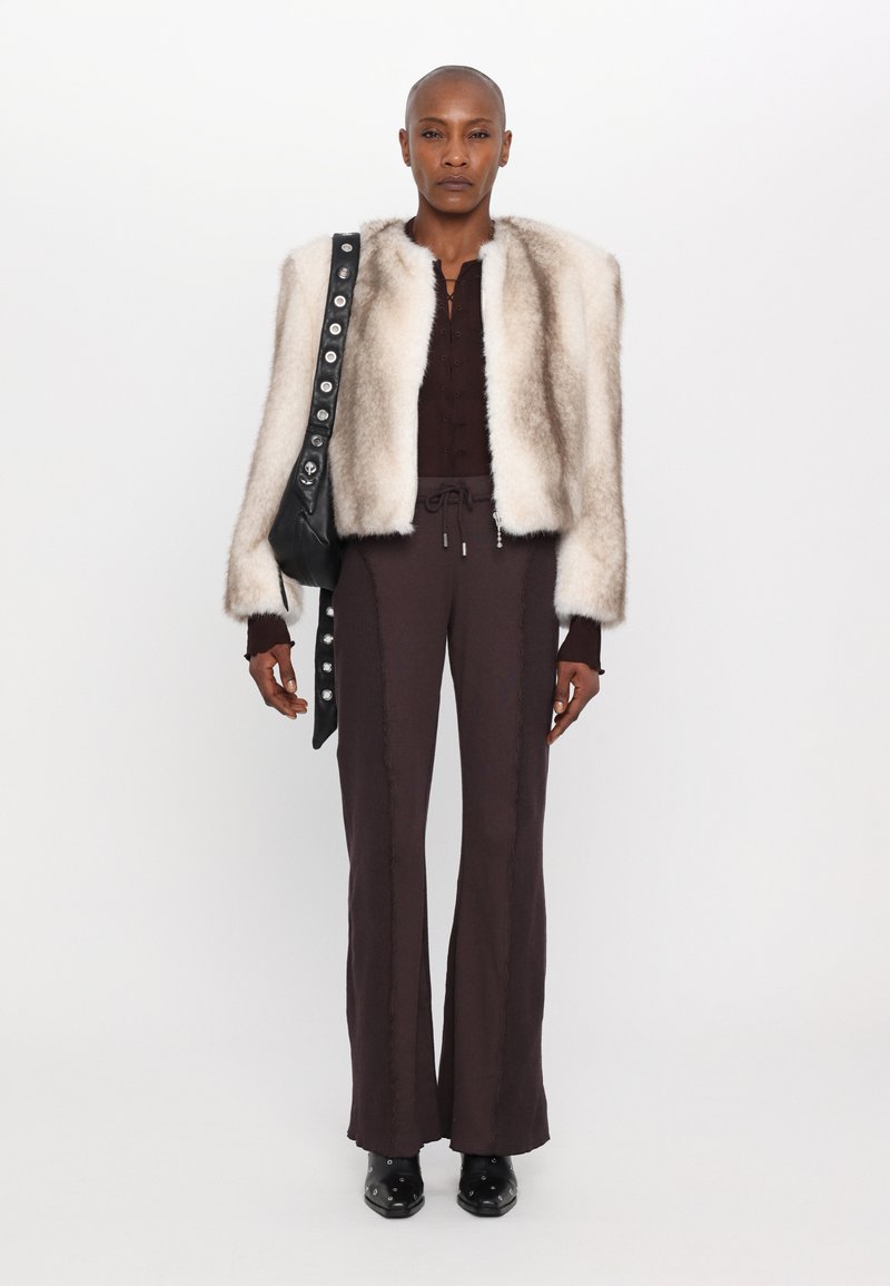 Chaqueta corta de piel sintética en crema, blusa de manga larga marrón con detalles de botones, pantalones marrones oscuros con costura lateral, bolso negro con botones plateados.