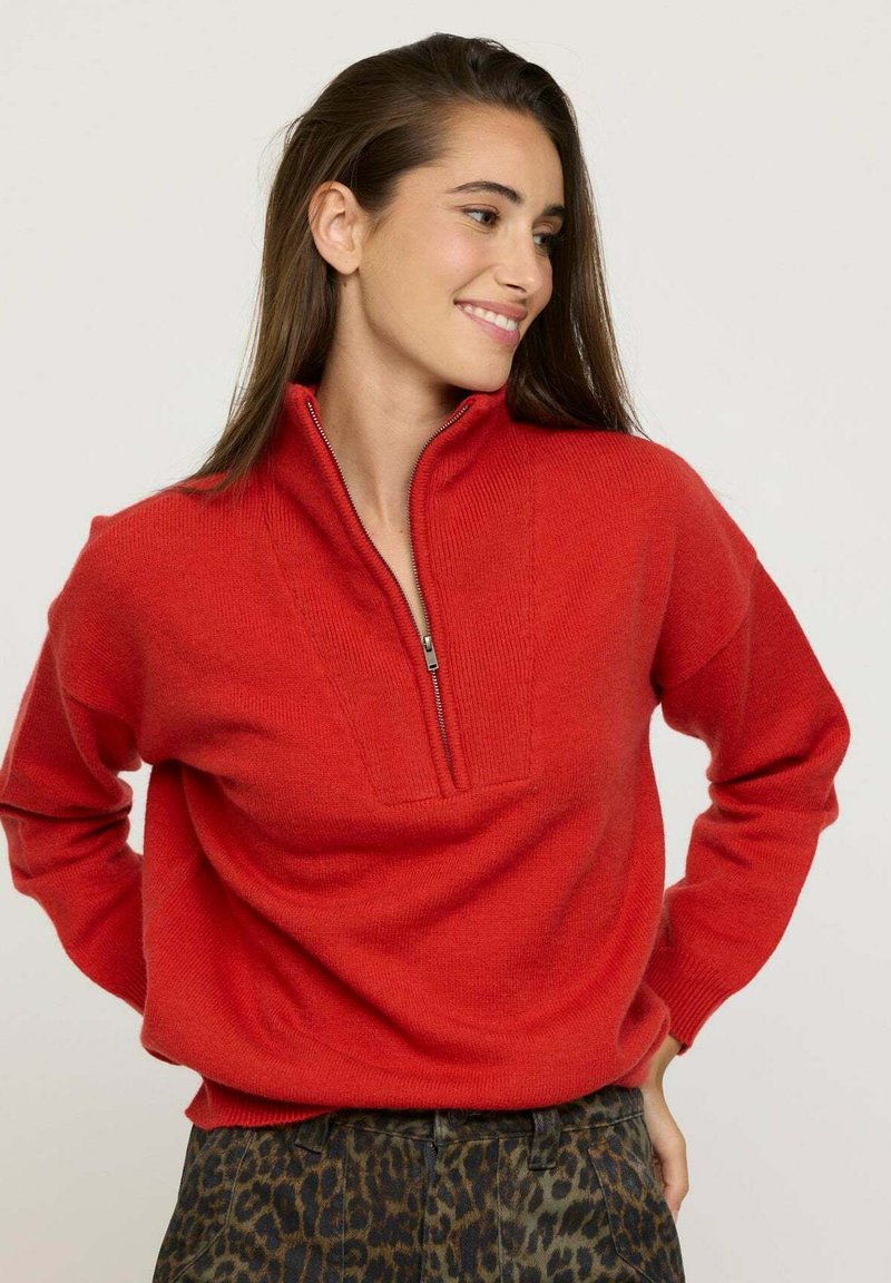Pull rouge avec col zippé quart, fabriqué en matériau doux. Présente une forme ajustée et des poignets côtelés, associé à un pantalon vert foncé à motifs.