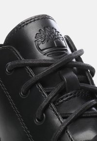 Czarne skórzane buty posiadają teksturowaną cholewkę z okrągłym noskiem, sznurowadłami oraz wytłoczonym logo drzewa na języku. Akcenty obejmują czarne szycie.