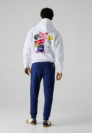 White hoodie with a colorful logo and patch design on the back, paired with navy joggers and multicolored sneakers.