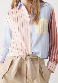 Multikleurige gestreepte bloes met roze, blauwe en rode secties; heeft een gele zak. Gepaard met beige hoge taillebroek.