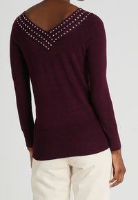 Burgundy long-sleeve sweater with a V-neck design, featuring silver studs along the neckline and a ribbed hem. Smooth texture.