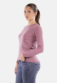 Maglione in maglia di un delicato viola chiaro con maniche lunghe, colletto rotondo e una sottile trama a coste, abbinato a jeans blu su uno sfondo neutro.