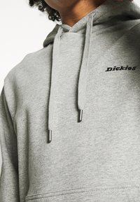 Felpa grigia realizzata in materiale morbido, con tasca frontale, cordini con punte in metallo e un logo ricamato discreto sul petto.