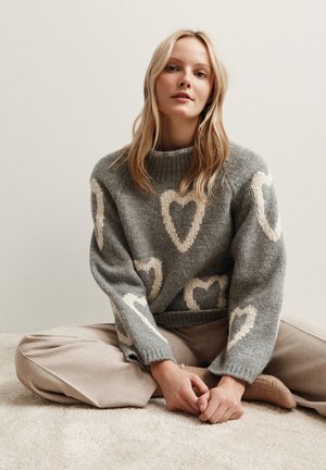 Pull en tricot gris avec des motifs de cœur crème, col rond et poignets côtelés, associé à un pantalon beige clair, assis sur une surface douce.