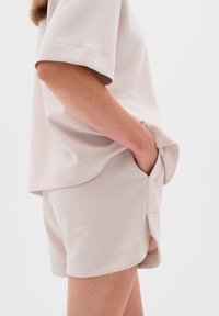 Světlý béžový outfit skládající se z krátkého rukávu a odpovídajících šortek. Tkanina se zdá být měkká s hladkou strukturou a bočními kapsami.