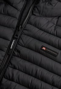 Sort puffjakke med quiltet struktur, med en frontlynlås og et Superdry-logo patch på brystet i rød og hvid.