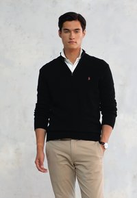 Schwarzer gestrickter Pullover mit einem Reißverschlusskragen, kombiniert mit einem weißen Hemd darunter. Das Modell trägt hellbeige Hosen und eine Uhr. Neutraler Hintergrund.