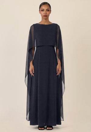 Frau in einem bodenlangen marineblauen Kleid mit transparenten Umhangärmeln, die aufrecht vor einem schlichten beigen Hintergrund steht.