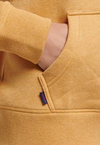Maglione giallo con polsini a coste e tasca frontale. Presenta un logo visibile in blu sulla tasca. Tessuto morbido e con una texture.