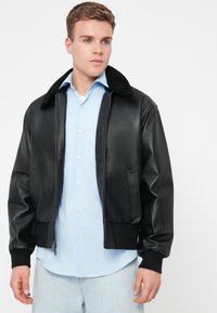 Fekete bőr bomberdzseki prémes gallérral, bordázott mandzsettákkal és cipzáras elülső zsebekkel, világoskék ing felett viselve.