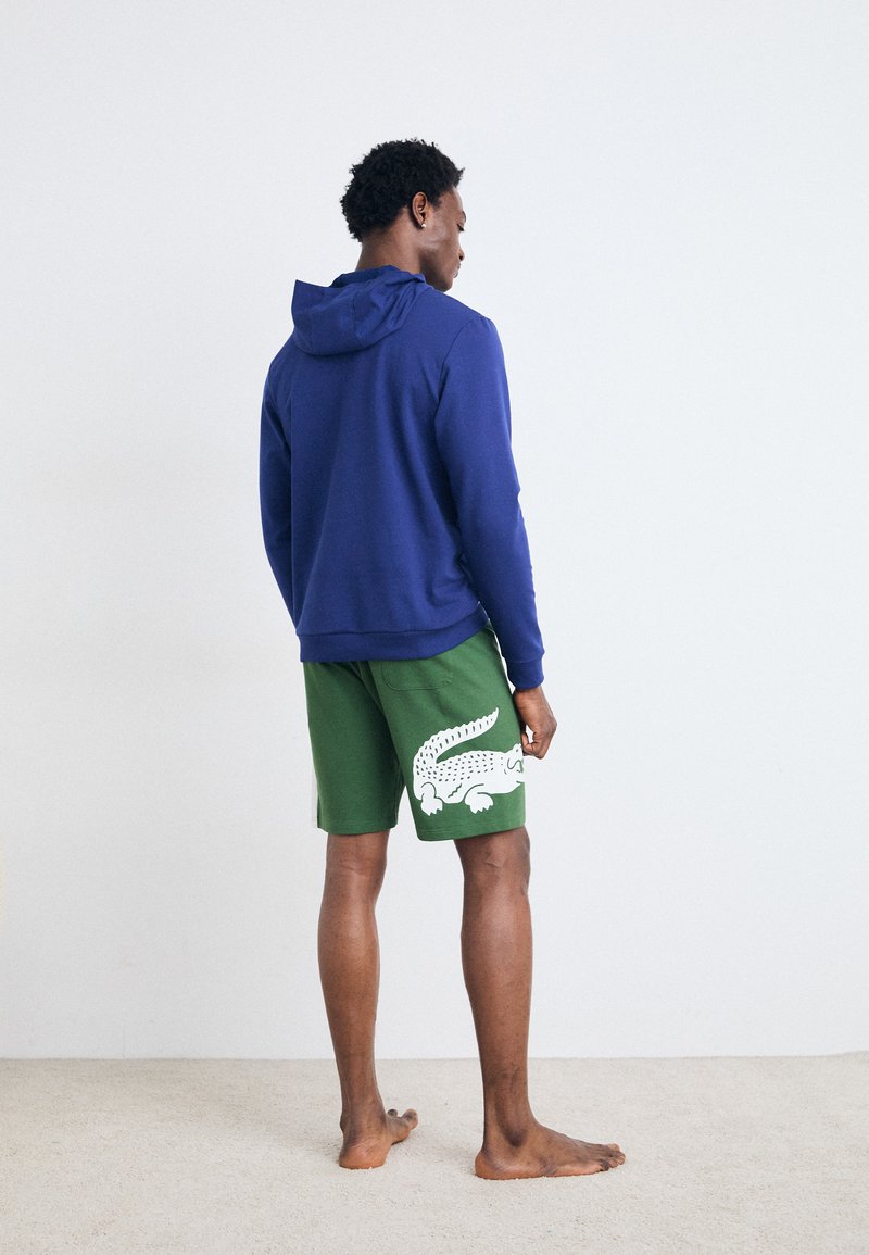 Blå hettegenser med snor, brukt over grønne shorts med et hvitt krokodillemotiv. Person som står med ryggen til, på et beige teppe.