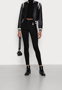 Veste bomber noire avec des accents à rayures blanches, associée à un jean skinny noir taille haute. Bottines noires et petit sac noir compris.